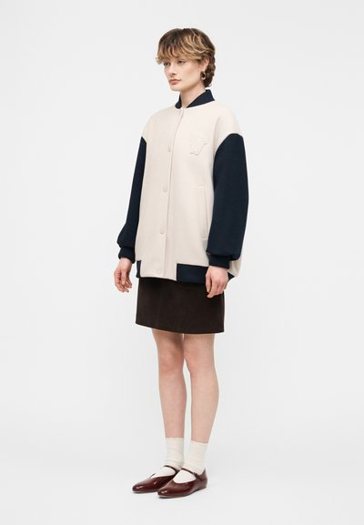 Νεαρό άτομο που στέκεται, φορώντας μπεζ και σκούρο μπλε μπουφάν τύπου bomber, μαύρη φούστα, λευκές κάλτσες και καφέ παπούτσια Mary Jane, μπροστά σε απλό φόντο.