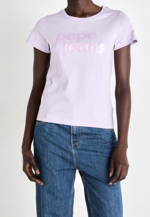 Persona de piel oscura con camiseta de Pepe Jeans color lila claro y vaqueros azules, de pie contra un fondo claro y liso.