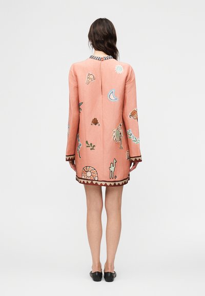 Γυναίκα που φοράει μακρυμάνικο ροζ φόρεμα με πολύχρωμα αφηρημένα σχέδια, στέκεται γυρισμένη προς τα πίσω, φορεί μαύρα φλατ παπούτσια σε απλό φόντο.