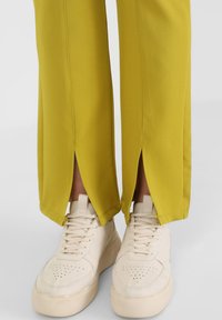 Żółte szerokie spodnie z rozcięciami po bokach u dołu, w połączeniu z jasnobeżowymi sneakersami z perforowanym wzorem i grubymi podeszwami.
