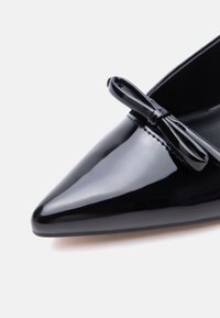 Mocassin plat en cuir verni noir à bout pointu avec un petit nœud sur le dessus, présenté en vue rapprochée et en angle.