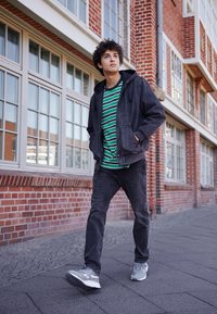 Jeune homme marchant sur un trottoir urbain à côté d'un immeuble en briques rouges avec de grandes fenêtres, portant une chemise rayée, une veste noire, un jean et des baskets grises.