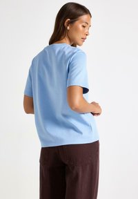Vrouw met lang haar, gekleed in een lichtblauw shirt met korte mouwen en donkerbruine broek, staand in een driekwart achteraanzicht tegen een witte achtergrond.