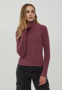 Maglione a collo alto aderente in materiale lavorato a coste e testurizzato. Il colore è un lampone smorzato, con maniche lunghe e colletto arrotolato.