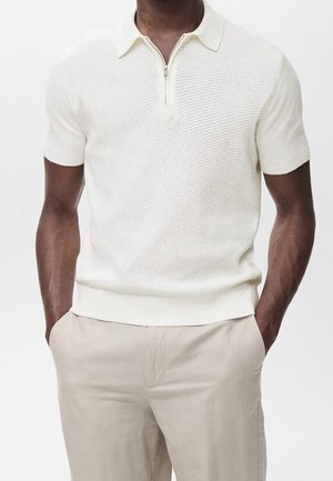 Mann trägt ein strukturiertes, weißes Kurzarm-Poloshirt mit Reißverschlusskragen und hellbeige Hose, Hände in den Taschen, vor einfarbigem Hintergrund.