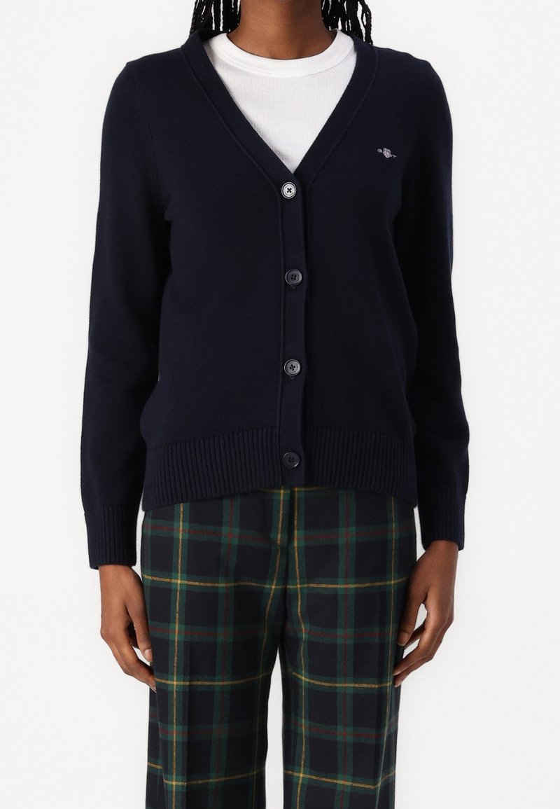 Personne portant un cardigan bleu marine boutonné sur un haut blanc et un pantalon à carreaux vert foncé avec des lignes jaunes et rouges.