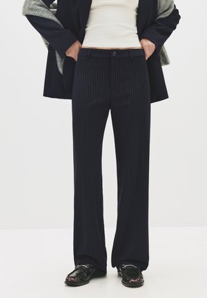 Person trägt schwarze Nadelstreifen-Weitbeinhose, schwarze Loafers, ein weißes Oberteil und einen dunkel gestreiften Blazer, der über graue Ärmel drapiert ist.