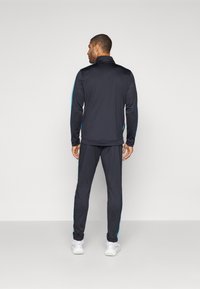 Survêtements de sport noirs en matériau doux, avec des rayures latérales bleu clair sur la veste et le pantalon, dotés d'un col zippé et d'une coupe slim.