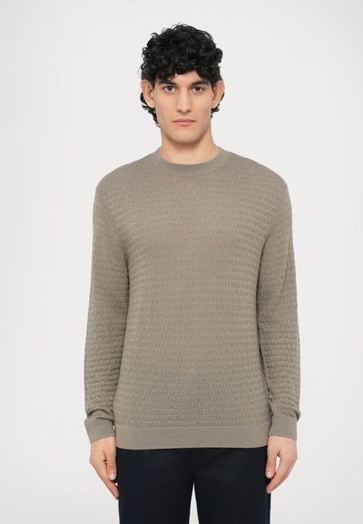 Pull vert clair texturé avec un col rond, présentant un motif de logo subtil et des poignets côtelés, associé à un pantalon foncé.