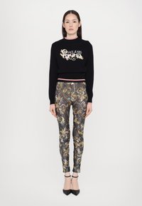 Svart cropped genser med "Versace Jeans Couture" logo; høy midje mønstrede leggings i gull, sølv og blå paljettdesign; svarte hæler.