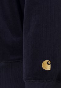 Marineblaues Cordstoffgewebe mit vertikalen Rippungen, ausgestattet mit einem goldbestickten Logo. Glatte Textur mit einer engen Passform am Handgelenk.