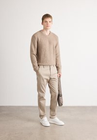 Beige strukturierter Strickpullover mit V-Ausschnitt, kombiniert mit hellbeigen Hosen und weißen Sneakern. Graue Umhängetasche mit langem Trageriemen.