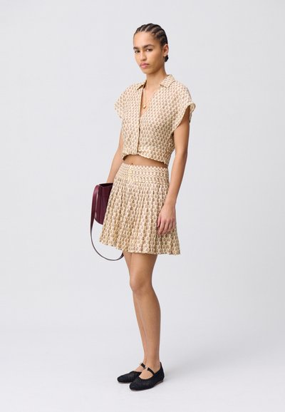 Crop top beige chiaro e gonna mini plissettata con motivo floreale; modella che tiene una borsa bordeaux; ballerine nere intrecciate; texture liscia.