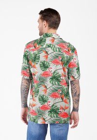 Mann mit Tattoos, der ein kurzärmliges Hemd mit grünem tropischem Blatt- und rosa Flamingo-Design trägt, kombiniert mit blauen Jeans, von hinten gezeigt.
