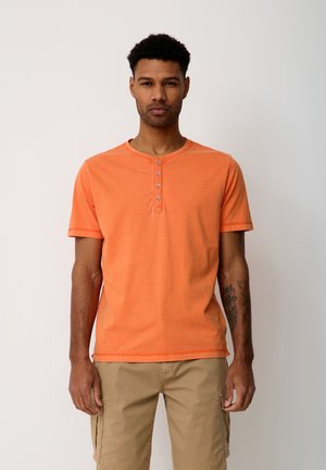 Mann mit kurzen lockigen Haaren, trägt ein orangefarbenes Henley-Shirt mit kurzen Ärmeln und beige Cargo-Hosen, steht vor einem schlichten hellen Hintergrund.