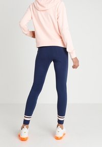 Person i marinblå leggings med rosa ränder vid anklarna, en ljusrosa hoodie och vita sneakers med orange sulor, står med en hand på höften.