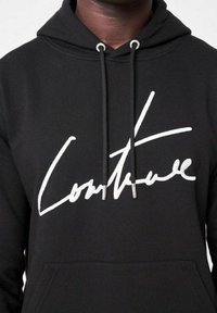 Svart hoodie i bomullsblandning med en stor vit kursiv text "Couture" framtill. Inkluderar snoddar med metallspetsar.