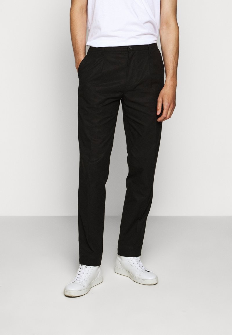 Muž v černých rovně střižených kalhotách, bílých teniskách a bílém tričku, stojící s rukama částečně v kapsách.