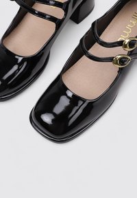 Zapatos de charol negros con punta cuadrada, pequeño tacón cuadrado y dos correas. Las hebillas en tono dorado realzan el diseño.