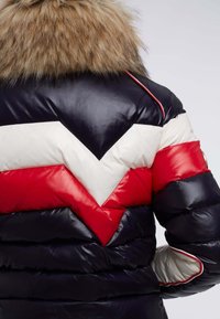 Veste matelassée avec un design en chevrons bleu marine, rouge et blanc, col en fausse fourrure marron et texture brillante.