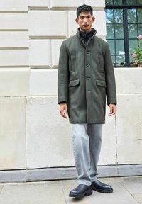 Grüner Wollmantel mit einem hohen Kragen, vier Fronttaschen und schwarzen Knöpfen, kombiniert mit hellgrauen Jeans und schwarzen klobigen Stiefeln.