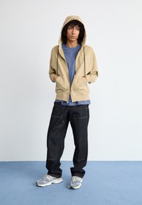 Pessoa jovem com cabelo encaracolado, vestindo uma hoodie bege, camisa azul, jeans escuros largos e tênis cinzas, de pé sobre um tapete azul contra um fundo branco.