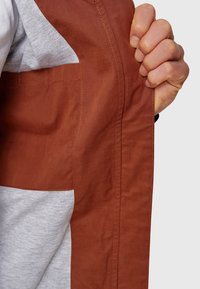 Main saisissant une veste marron ouverte révélant la poche intérieure et une chemise grise en dessous sur un fond neutre.