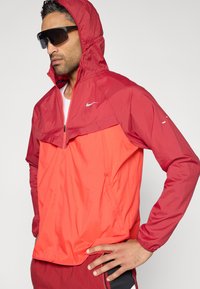 Ελαφρύ κόκκινο και πορτοκαλί μπουφάν με κουκούλα, μερικό φερμουάρ και λογότυπο Nike. Υφασμάτινη υφή με σχεδιασμό σε μπλοκ χρωμάτων και ελαστικά μανίκια.