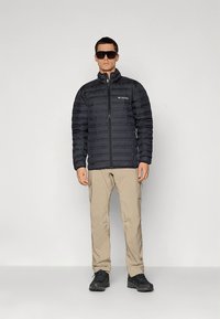 Svart vattert jakke med horisontalt quiltet design, glidelåslukking og logo. Beige cargobukser og svarte sko fullfører antrekket.