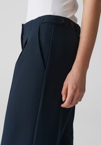 Op maat gemaakte marineblauwe pantalons met plooien, met een soepel textuur, dubbele knoopsluiting en een aangesloten tailleband. Hand aan de zijkant gepositioneerd.