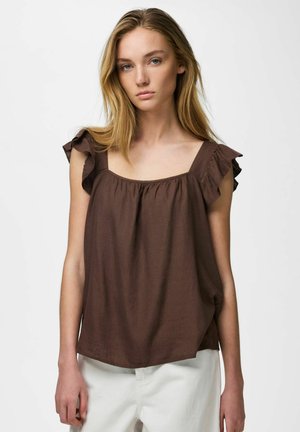 Ung kvinne med langt blondt hår iført en brun ermeløs bluse med volang på skuldrene og hvite bukser, stående foran en ensfarget bakgrunn.