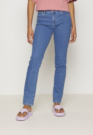 Persoon die lichtblauwe slim-fit jeans, een roze top en lavendelkleurige open-toe platformsandalen draagt, staand tegen een witte achtergrond.