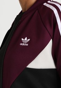 Vinröd och svart zip-jacka med vita detaljer, prydd med tre vita ränder på axeln och en broderad Adidas-logotyp.