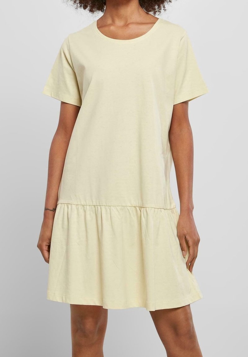 Femme portant une robe jaune clair à manches courtes, longueur genou, avec une jupe froncée sur un fond gris uni.