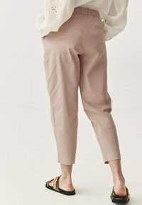 Light pink, cropped pants with an elastic waistband and two back pockets. Worn with flat black sandals featuring a gold buckle detail.