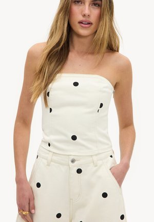 Femme portant un haut blanc sans bretelles et un pantalon assorti à gros pois noirs, une main dans la poche, cheveux blonds longs lâchés.