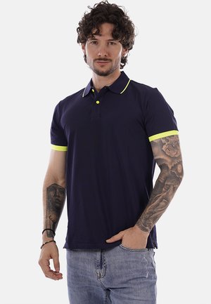 Uomo con capelli ricci che indossa una polo blu scuro con bordi gialli e jeans azzurri, mostrando tatuaggi su entrambi gli avambracci.