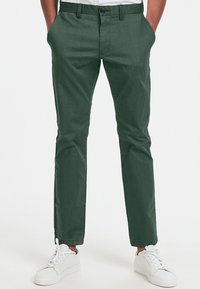 Gröna slim-fit chinos med en slät textur, som har en knappstängning, snedställda fickor och en avsmalnande bendesign, kombinerat med vita sneakers.
