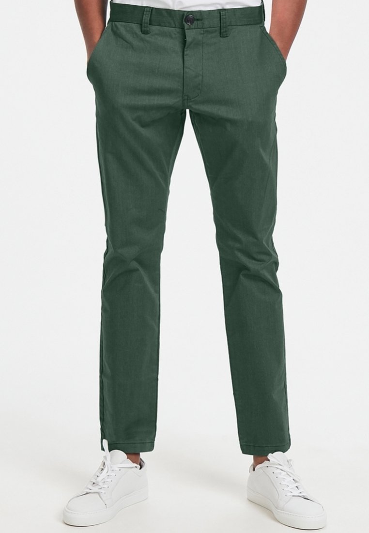Gröna slim-fit chinos med en slät textur, som har en knappstängning, snedställda fickor och en avsmalnande bendesign, kombinerat med vita sneakers.