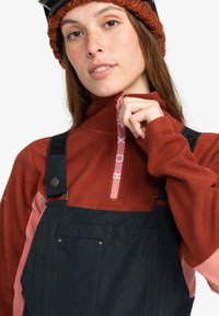 Sudadera de forro polar en óxido con media cremallera y acentos en rosa, combinada con un overall negro con tirantes ajustables y bolsillos.