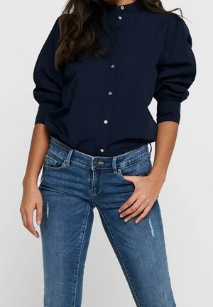 Femme portant un chemisier bleu marine à boutons avec manches bouffantes et un jean skinny bleu déchiré, debout avec une main dans la poche.