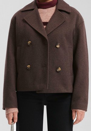 Bruine dubbelgeknoopte wollen jas gedragen over een roze coltrui, gecombineerd met een zwarte broek, een persoon die een zilveren kettingaccessoire vasthoudt.