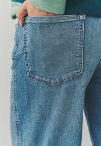 Gai zila denim bikses ar lielu, taisnstūrveida aizmugurējo kabatu. Audumam ir gluda tekstūra, un redzamā šuvju akcenti izceļ malas.