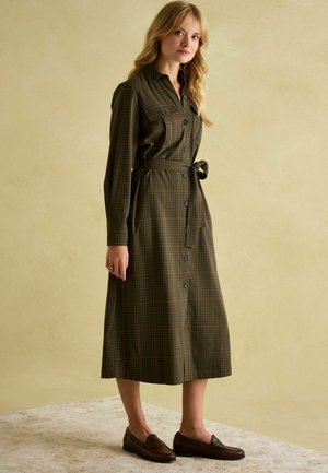 Langärmliges, geknöpftes Kleid aus dunklem, kariertem Stoff mit gebundenem Taillenbund, seitlichen Taschen und knöchellanger Passform. Getragen mit braunen Loafers.