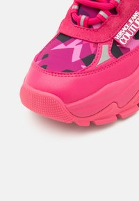 Lyserød sneaker med tekstureret gummisål, abstrakt geometrisk mønster, ruskindsdetaljer, reflekterende elementer og "Versace Jeans Couture" logo.
