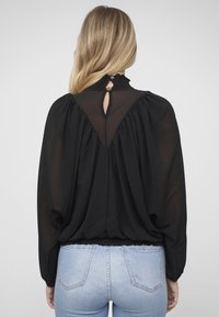 Schwarze, durchsichtige Bluse mit gerafftem Kragen und Schlitz am Rücken. Lange Ärmel und gerafftes Design mit elastischem Bund. Kombiniert mit blauen Jeans.