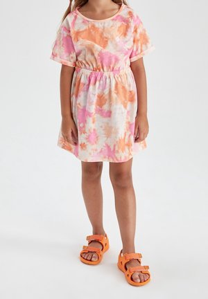 Barn iført kortærmet, pink og orange batikfarvet kjole med elastisk talje og lyse orange sandaler, stående på hvid baggrund.