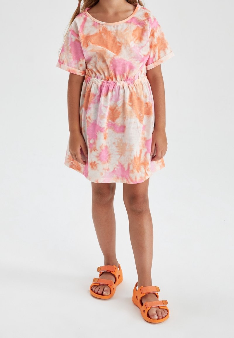 Enfant portant une robe à manches courtes tie-dye rose et orange avec une taille élastique et des sandales orange vif, debout sur un fond blanc.