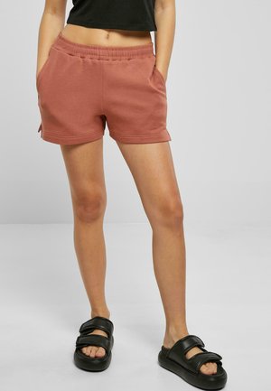 Rust-colored cotton shorts with an elastic waistband, side pockets, and a relaxed fit. Paired with black slide sandals.