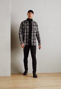 Camicia a quadri bianco e nero sopra un dolcevita nero, abbinata a pantaloni neri slim-fit e scarpe nere, su uno sfondo neutro.